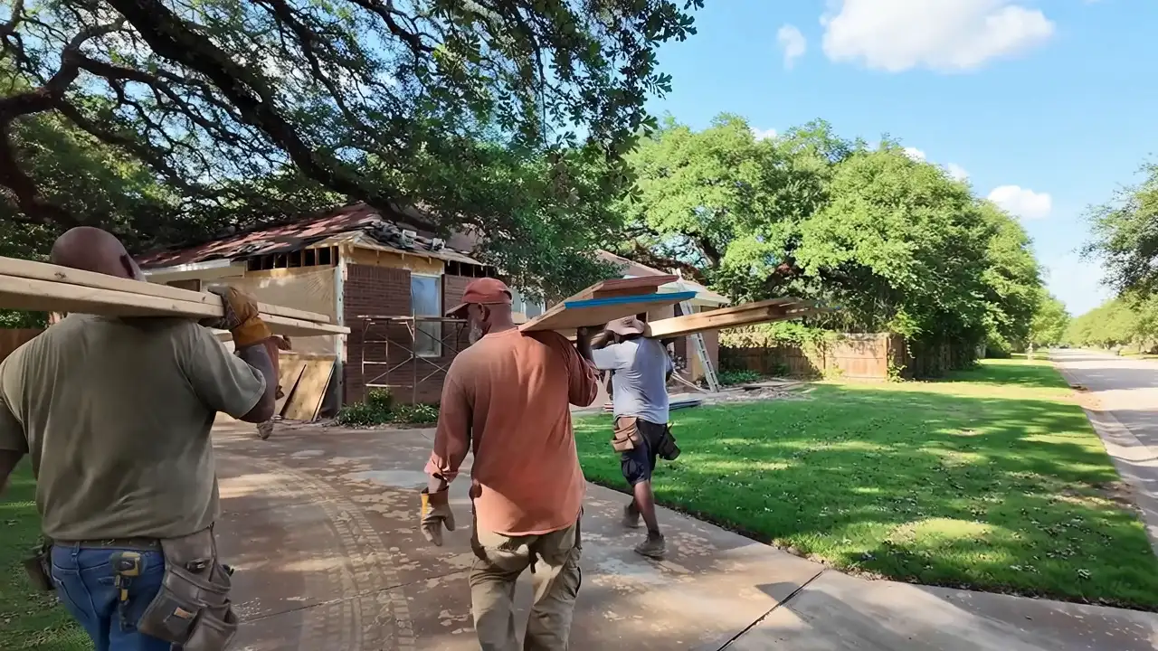 Workers seen from behind carrying lumber toward a brick house under restoration